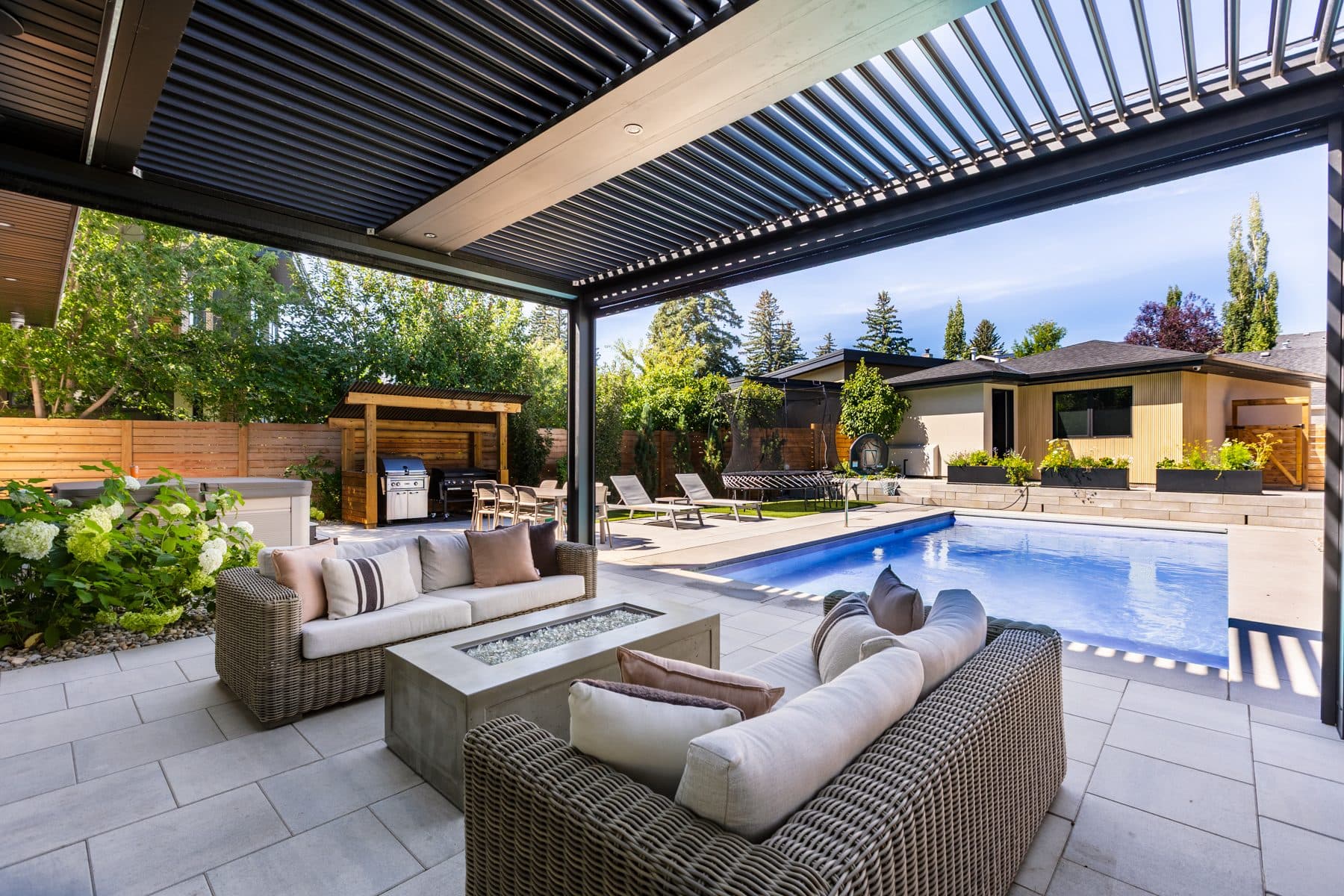 An outdoor seating group around a fire table, sheltered by a louvred pergola.